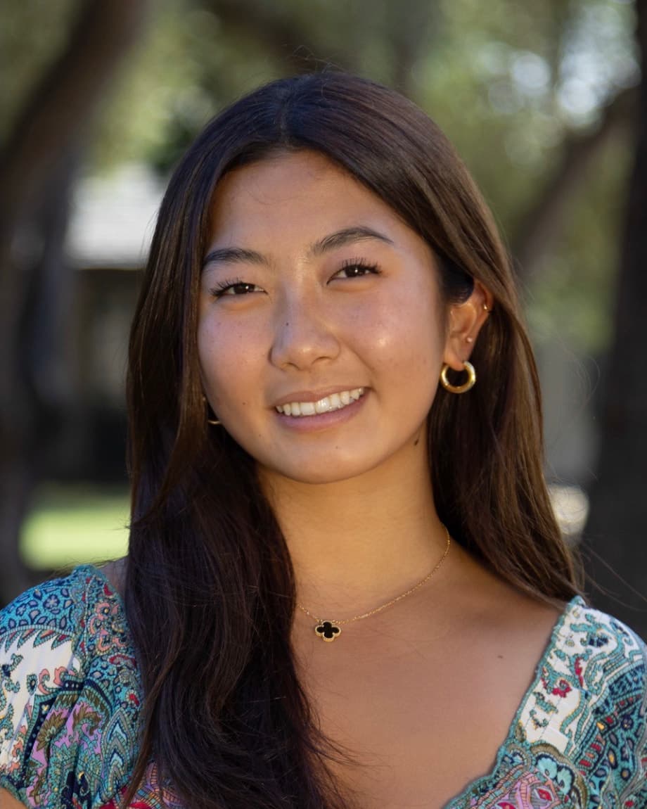 Alyna outdoors in a patterned blue top, smiling at the camera, trees blurred behind her
